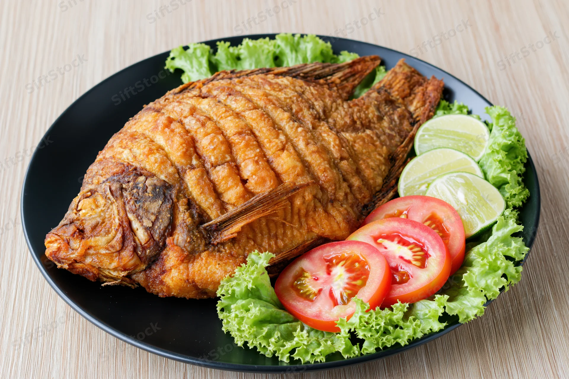 Whole deep-fried tilapia with crispy golden-brown skin served on a black plate with lettuce, tomato, and lime slices