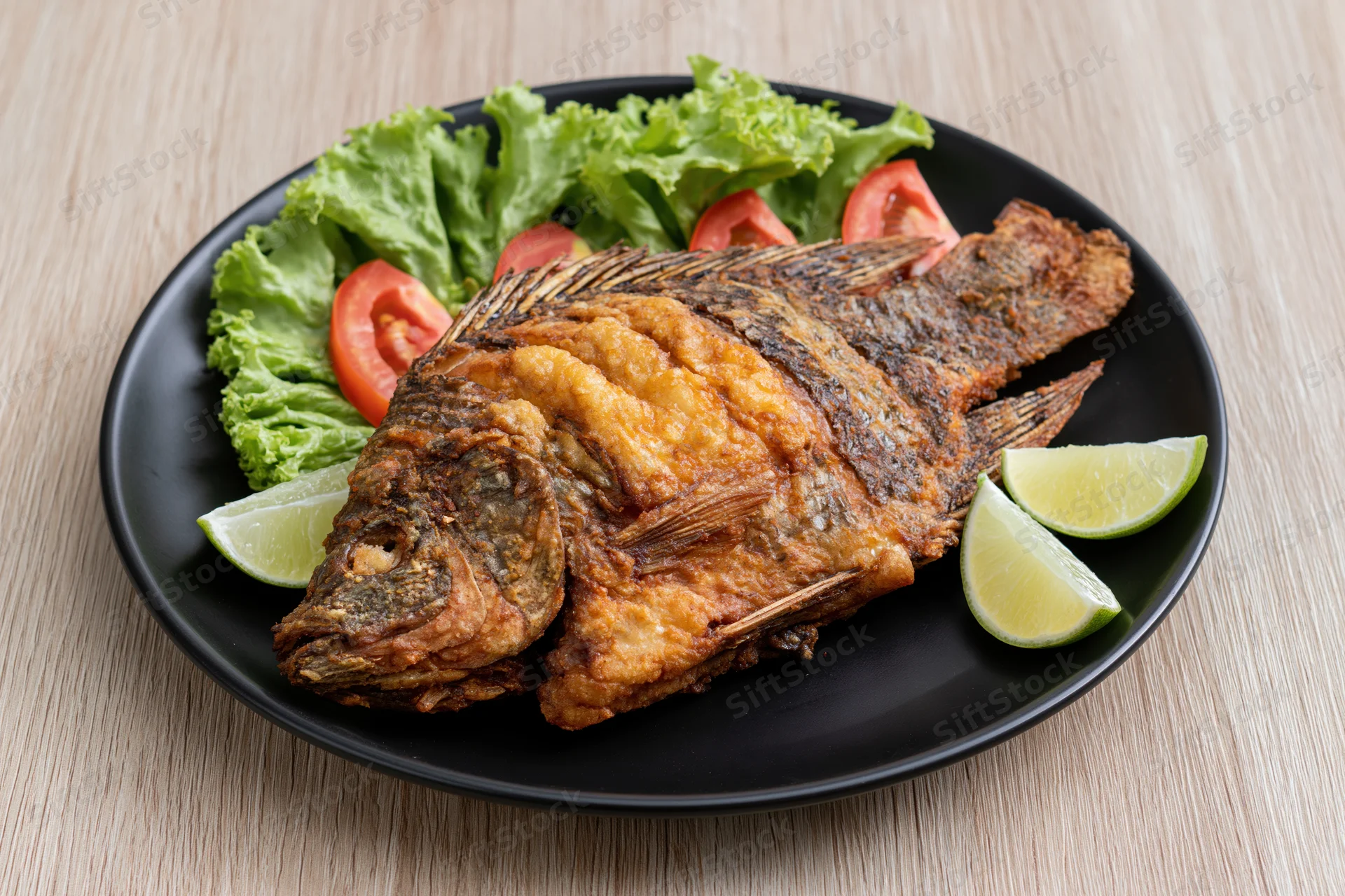 Whole fried tilapia with crispy skin served on black plate with lettuce tomato and lime wedges