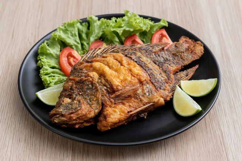 Whole fried tilapia with crispy skin served on black plate with lettuce tomato and lime wedges