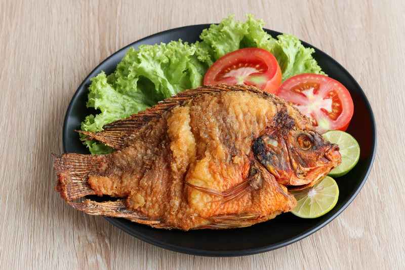 Whole fried tilapia with crispy skin served on black plate with lettuce tomato and lime slices