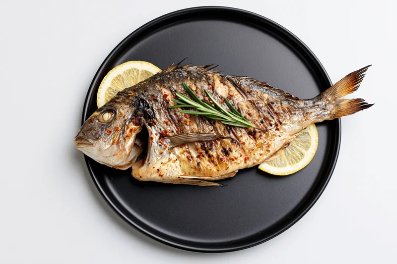Whole grilled sea bream on a black ceramic plate with lemon and rosemary garnish
