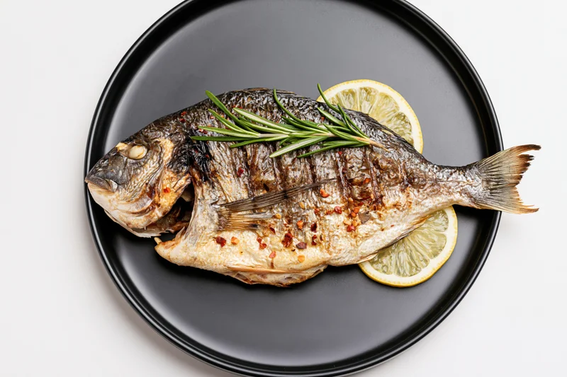 Whole grilled sea bream on a black ceramic plate with lemon and rosemary garnish