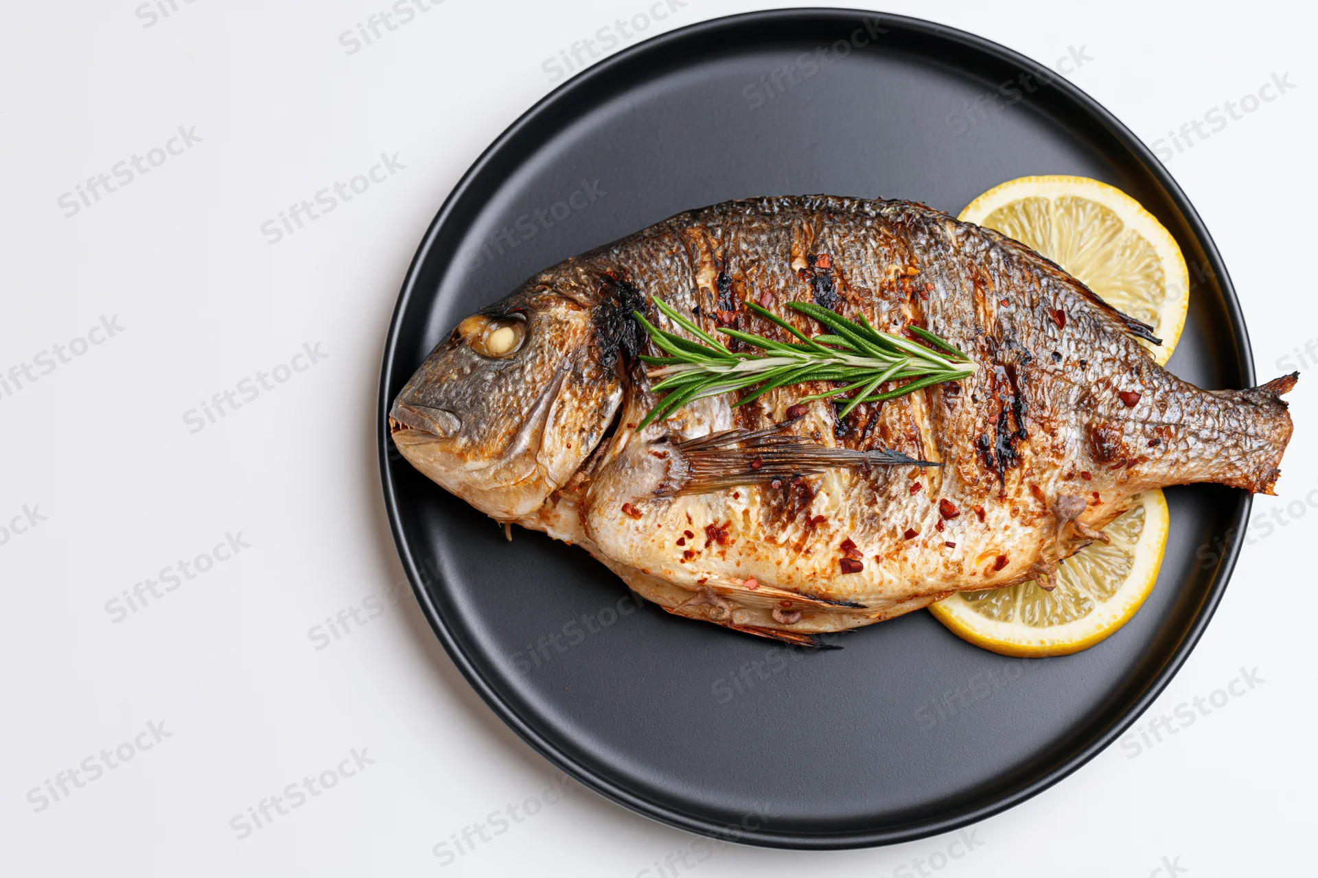 Whole grilled sea bream on a black ceramic plate with lemon and rosemary garnish, top view on white background