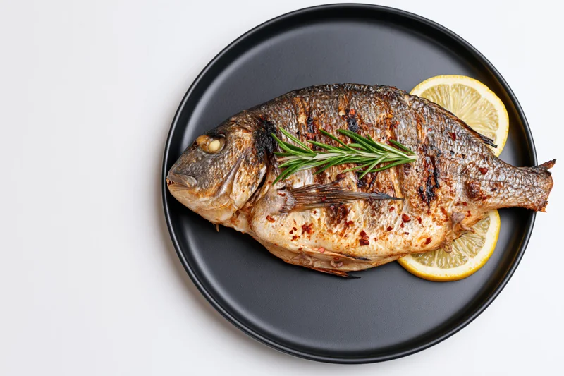 Whole grilled sea bream on a black ceramic plate with lemon and rosemary garnish, top view on white background