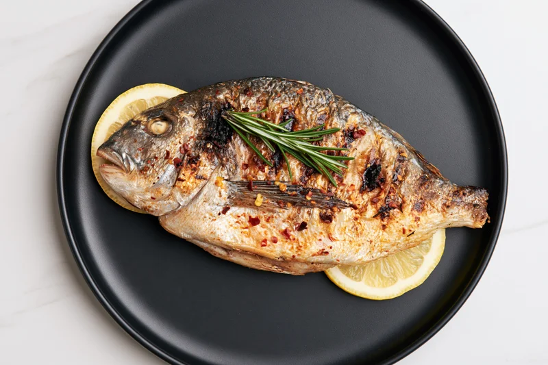 Whole grilled sea bream on a black ceramic plate with lemon and rosemary garnish