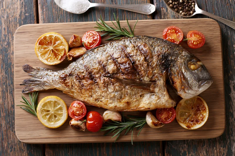 Whole grilled sea bream on a wooden cutting board with lemon, tomatoes, rosemary and garlic