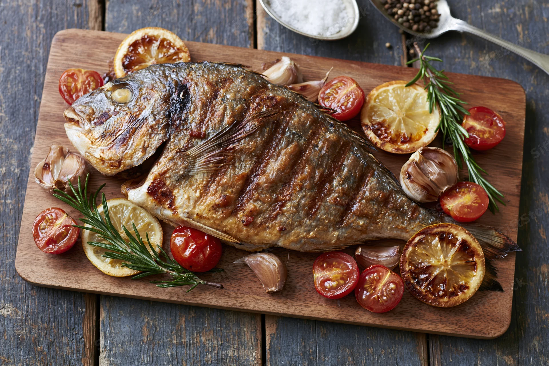 Whole grilled sea bream on a wooden cutting board with tomatoes, lemon, rosemary and garlic