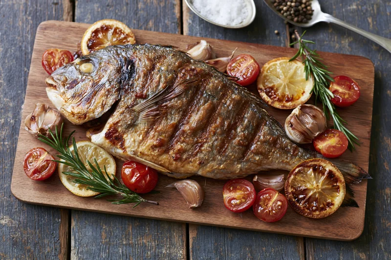 Whole grilled sea bream on a wooden cutting board with tomatoes, lemon, rosemary and garlic