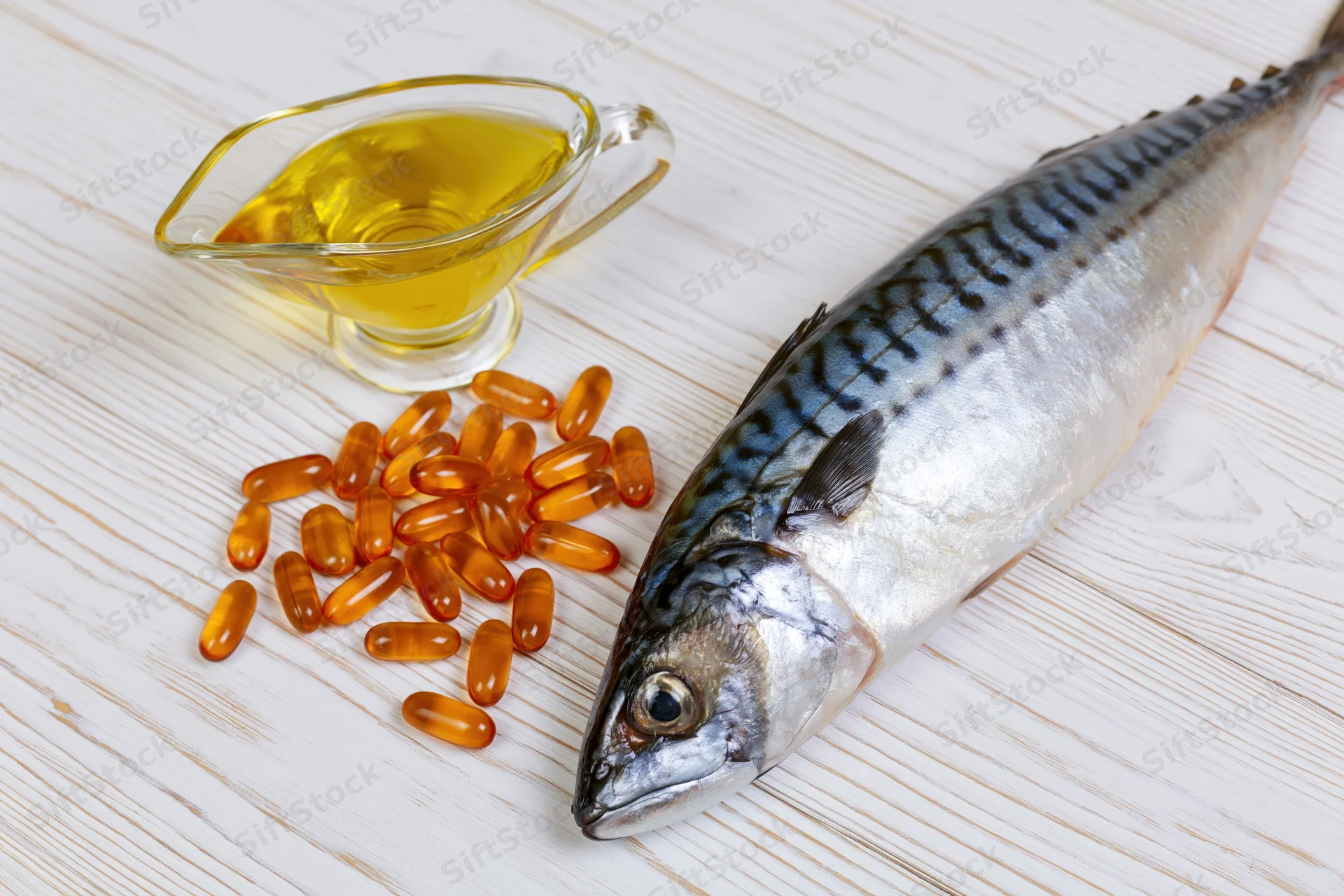 Whole raw mackerel fish lying beside fish oil capsules and a glass pitcher of oil on a white-painted wooden surface