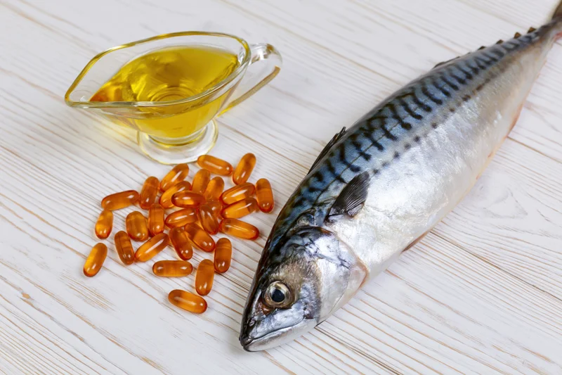 Whole raw mackerel fish lying beside fish oil capsules and a glass pitcher of oil on a white-painted wooden surface