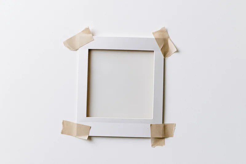 Blank polaroid frame taped to white surface with beige paper tape, overhead view