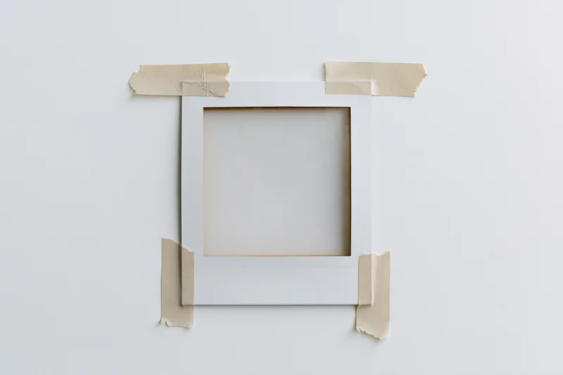 Blank polaroid frame taped to white surface with beige adhesive tape, centered, flat lay view