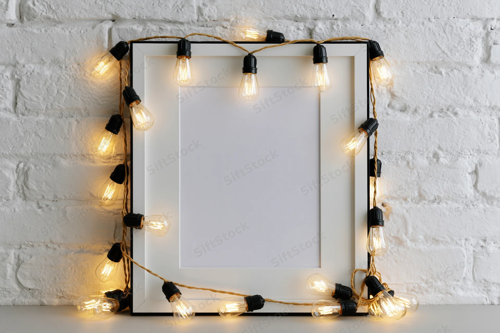 Blank white picture frame with black border leaning against white brick wall, decorated with glowing string lights