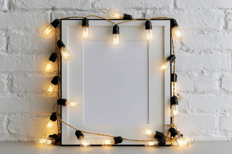 Blank white picture frame with black border leaning against white brick wall, decorated with glowing string lights