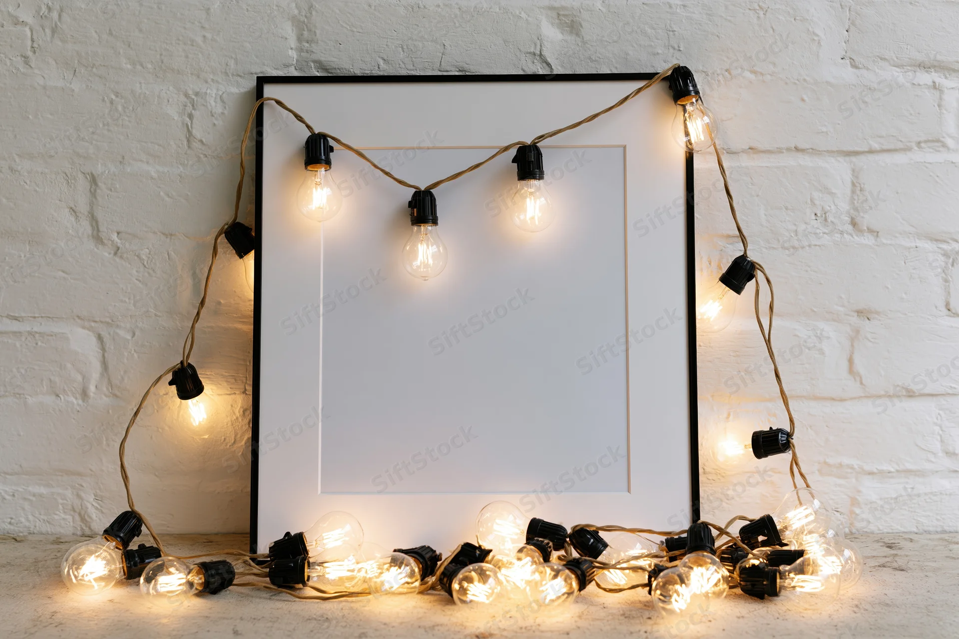 Blank white picture frame with black border leaning against brick wall, surrounded by glowing string lights