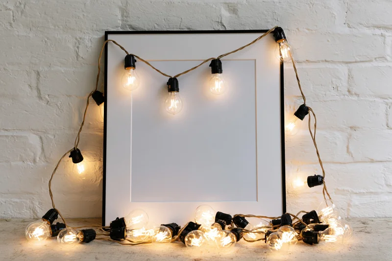 Blank white picture frame with black border leaning against brick wall, surrounded by glowing string lights