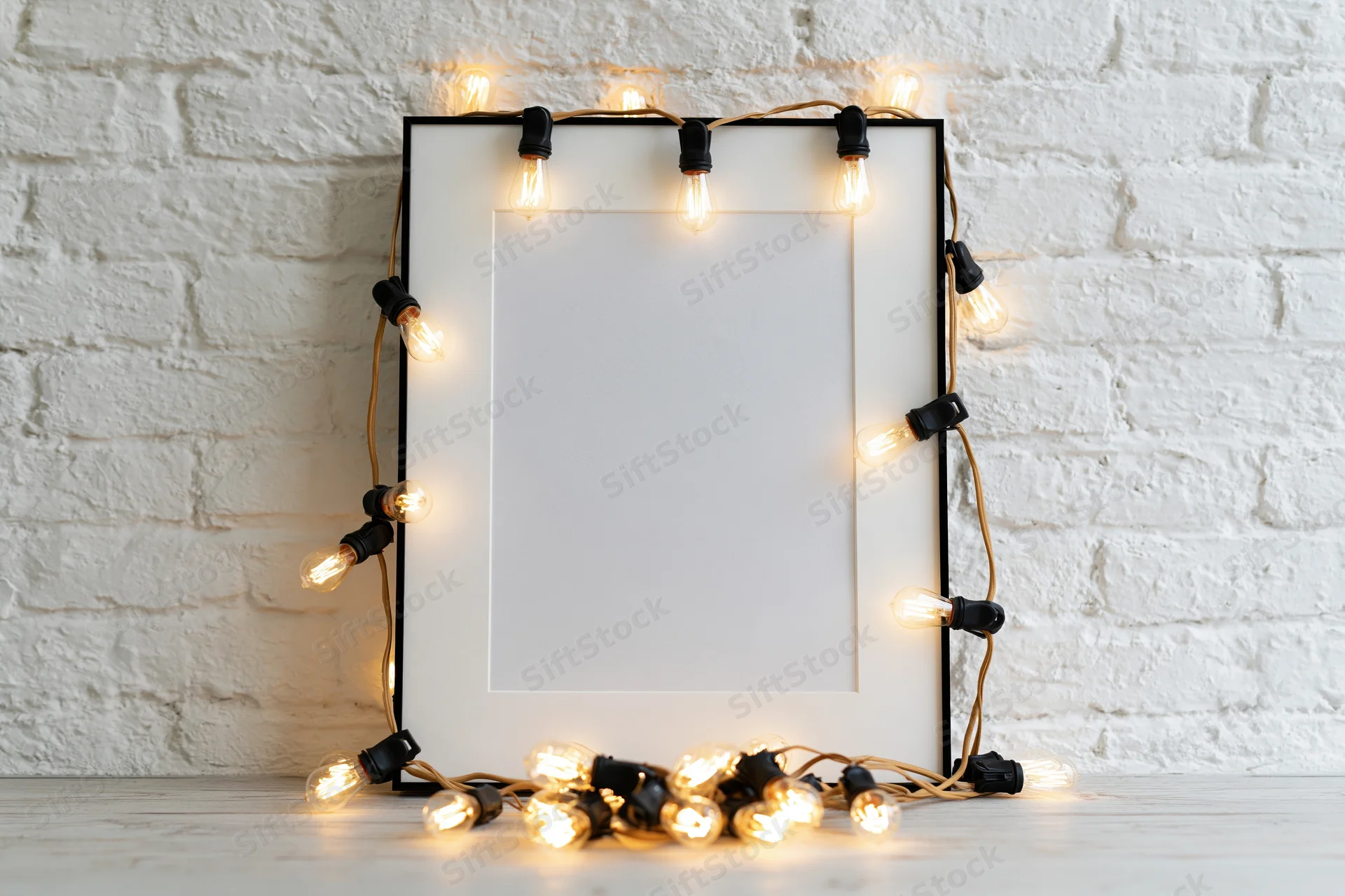 Blank white picture frame with black border decorated with string lights leaning against white brick wall on wooden floor