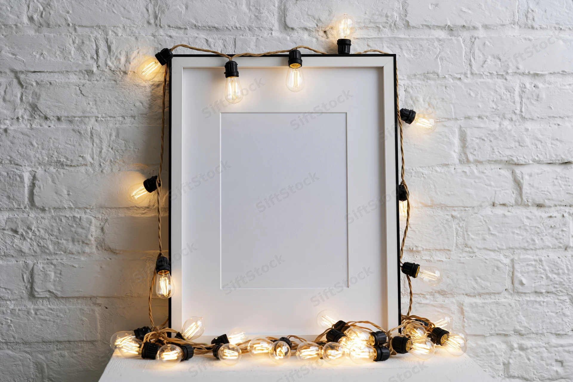 Blank white picture frame with black border leaning against painted brick wall, surrounded by warm string lights on table