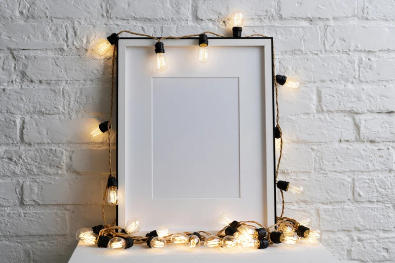 Blank white picture frame with black border leaning against painted brick wall, surrounded by warm string lights on table