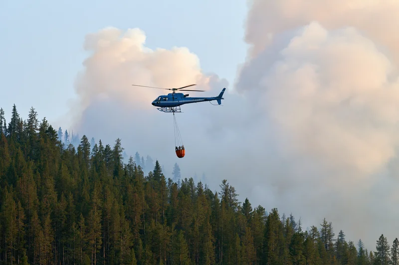 Blue helicopter carrying orange water bucket over smoky forest during firefighting operation