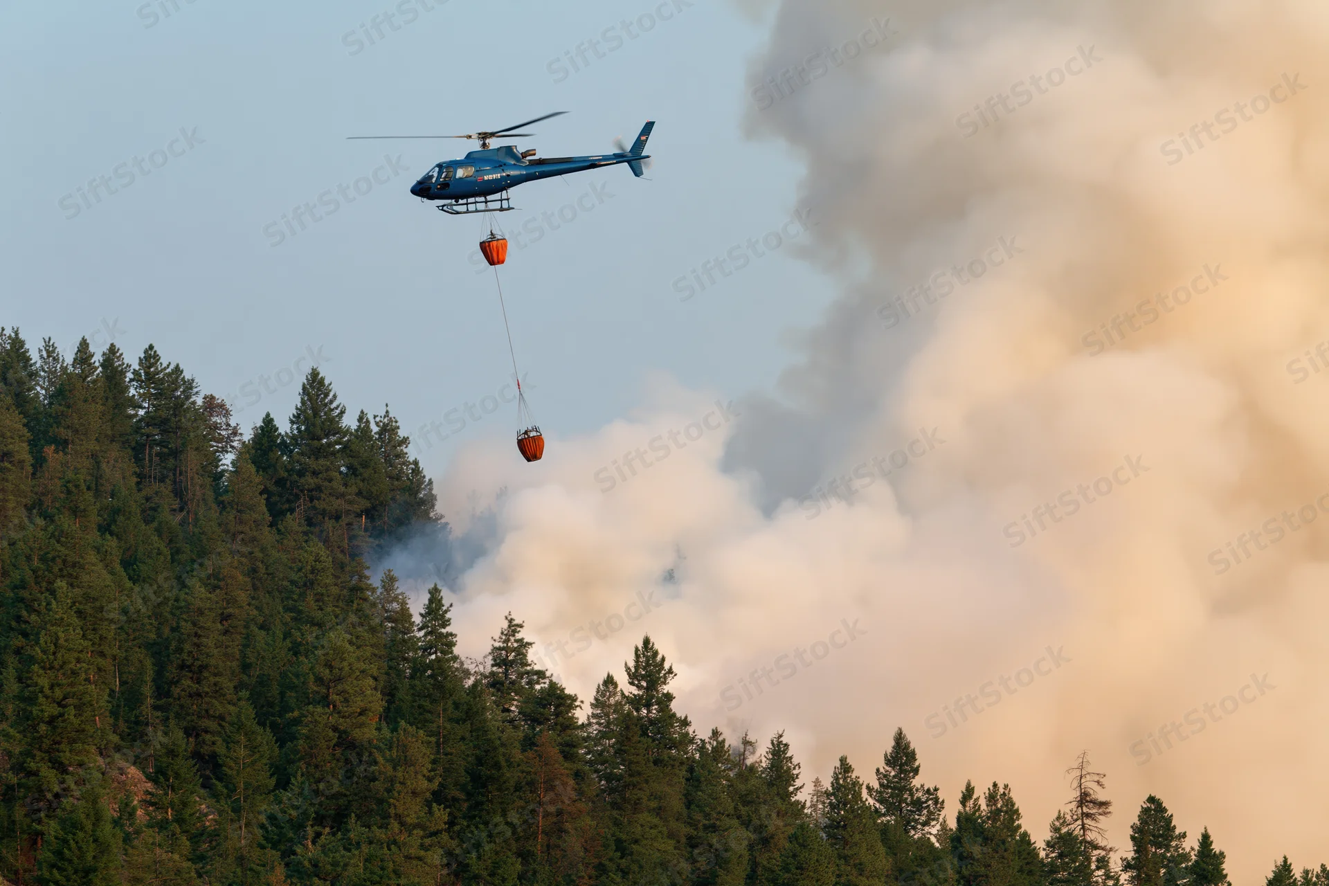 Blue helicopter carrying orange water bucket over smoky forest during wildfire firefighting operation