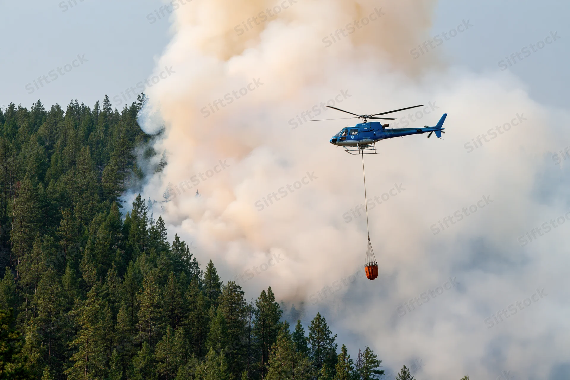 Blue helicopter carrying orange water bucket over smoky forest during wildfire suppression operation
