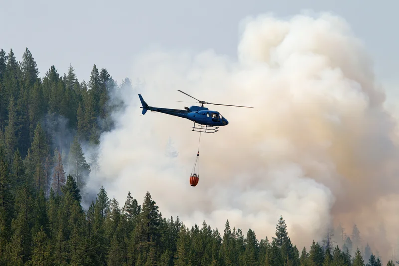 Blue helicopter carrying orange water bucket over smoky forest during wildfire suppression operation