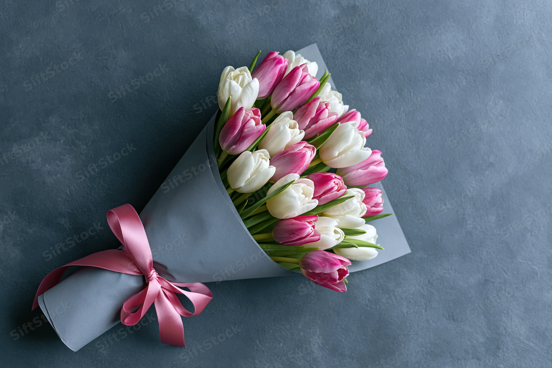 Bouquet of pink and white tulips wrapped in grey paper with pink ribbon on dark surface
