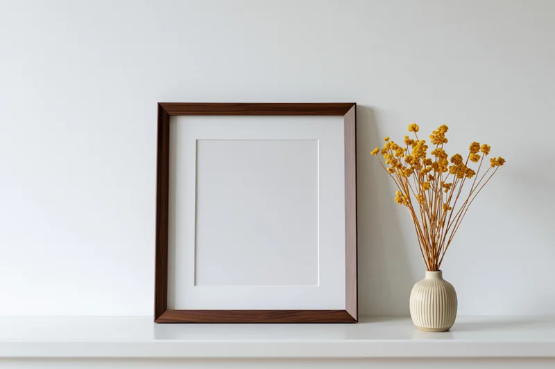 Dark walnut wood picture frame with white mat leaning on white shelf beside a ceramic vase holding dried yellow flowers