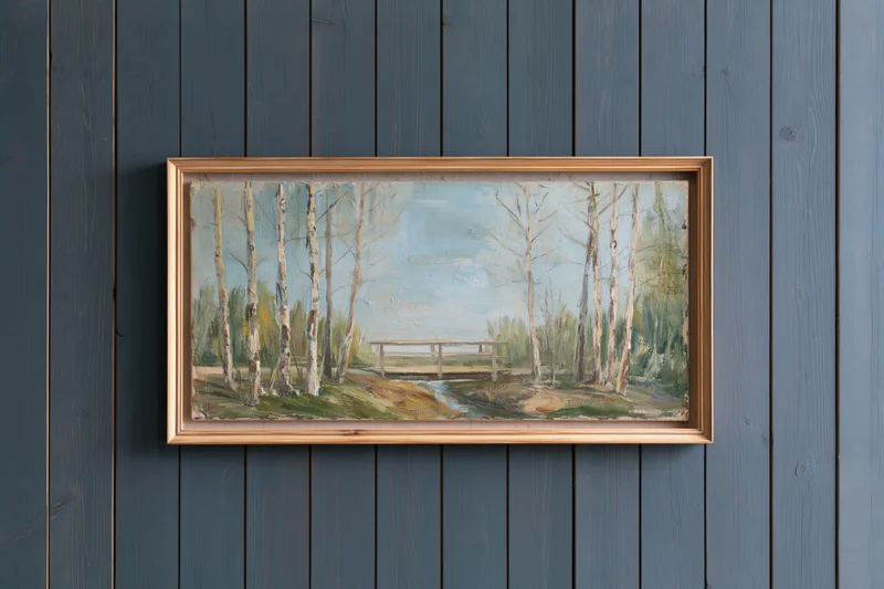 Framed oil painting of birch trees and a wooden bridge over a stream hanging on a dark grey wooden wall