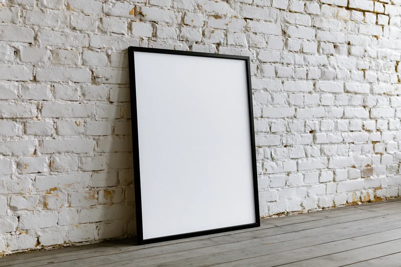Black-framed blank poster leaning against white brick wall in studio space