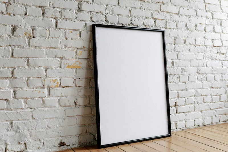 Black-framed blank poster leaning against white brick wall on wooden floor