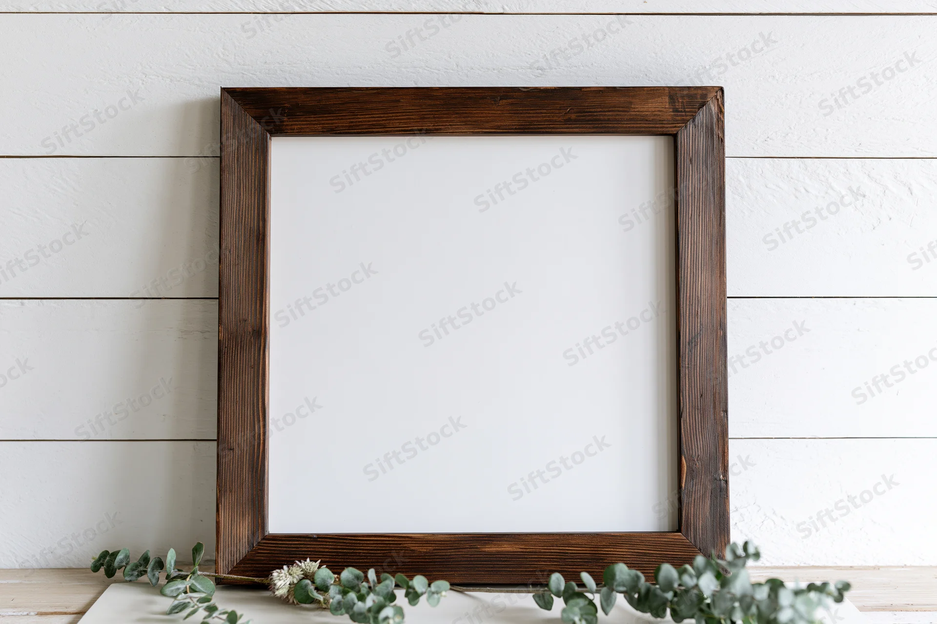 Dark stained wooden frame with blank white interior leaning on white shiplap wall, surrounded by eucalyptus greenery on wooden shelf