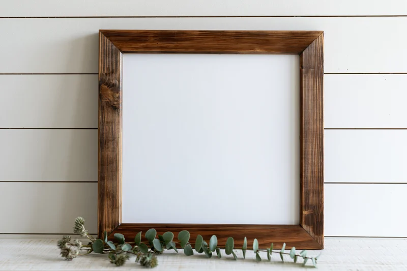 Dark stained wooden frame with blank white interior and eucalyptus branch on shiplap wall