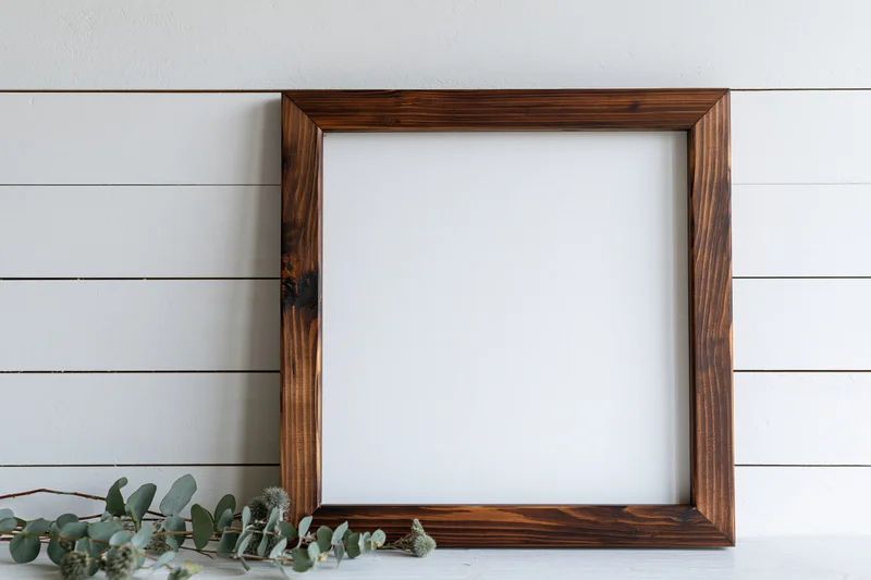 Dark stained wooden frame with blank white interior on white shiplap wall, eucalyptus branch beside it