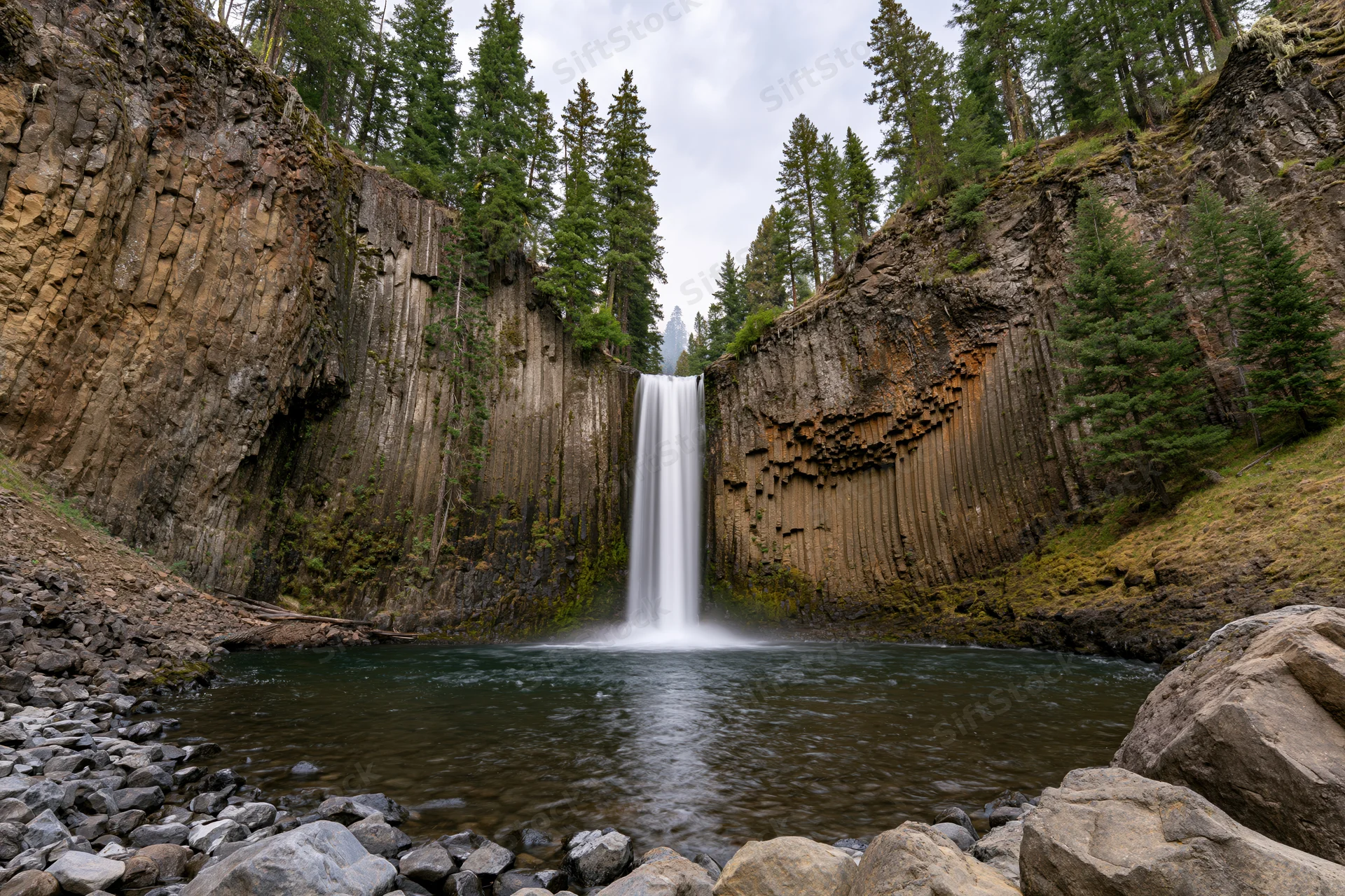 Waterfall dropping into a calm pool at the base of a rocky canyon surrounded by pine trees