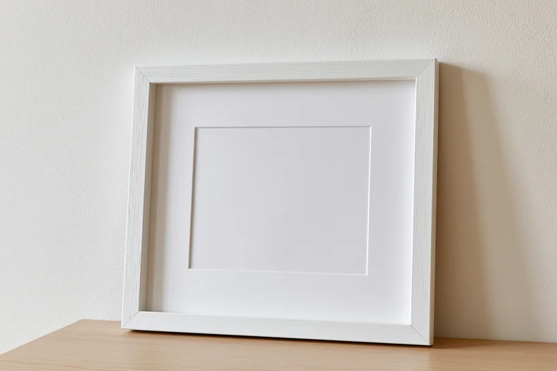 White picture frame with white mat leaning against a wall on a wooden surface