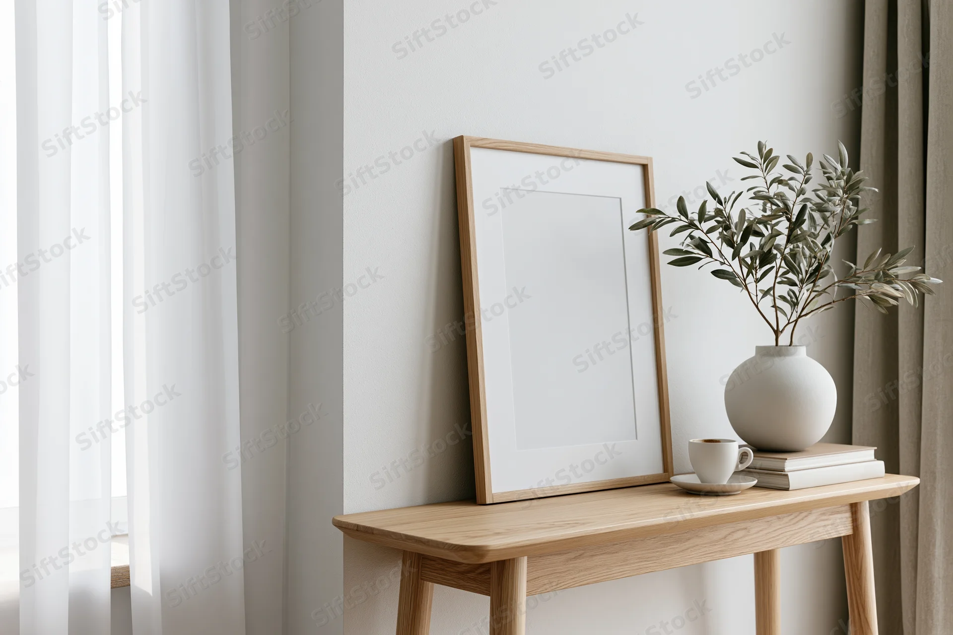 Wooden frame leaning against white wall on console table with vase of olive branches, books, and cup, near window with sheer curtain