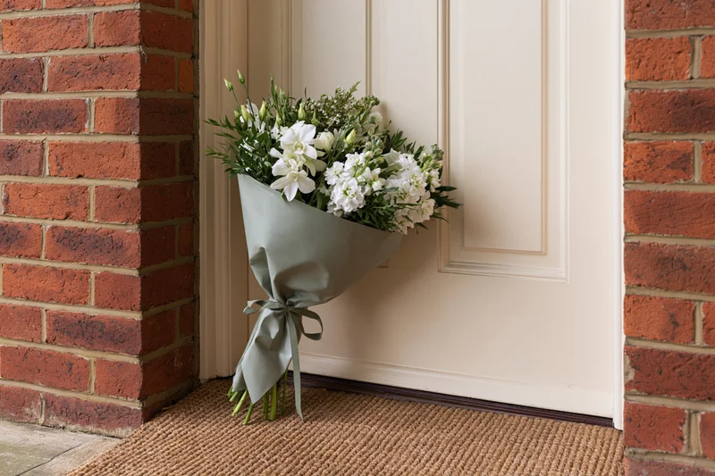 Wrapped bouquet of white flowers and green foliage leaning against a cream door on a brick porch