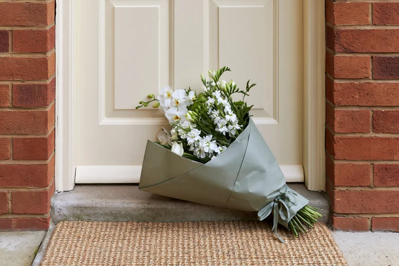 Wrapped bouquet of white flowers and green foliage resting on doorstep in front of cream door flanked by red brick walls