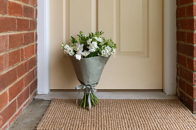 Wrapped bouquet of white flowers and green foliage resting on a woven mat at a front door entrance
