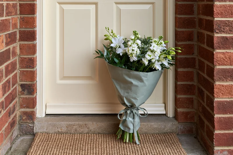 Wrapped bouquet of white flowers and green foliage resting on doormat in front of cream door flanked by red brick walls