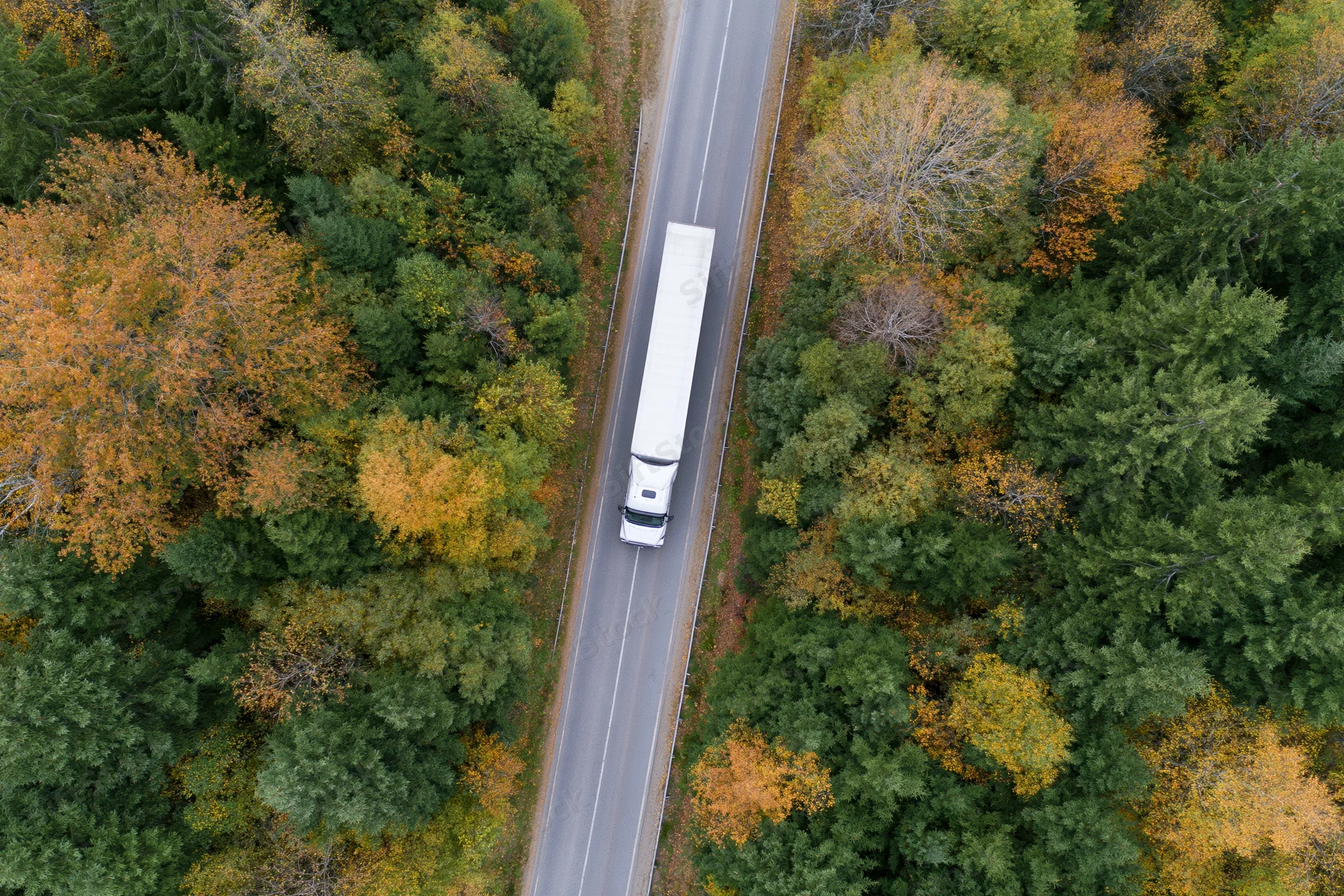 White semi-truck driving on a highway through autumn forest from aerial top-down view