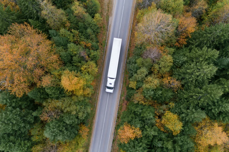 White semi-truck driving on a highway through autumn forest from aerial top-down view