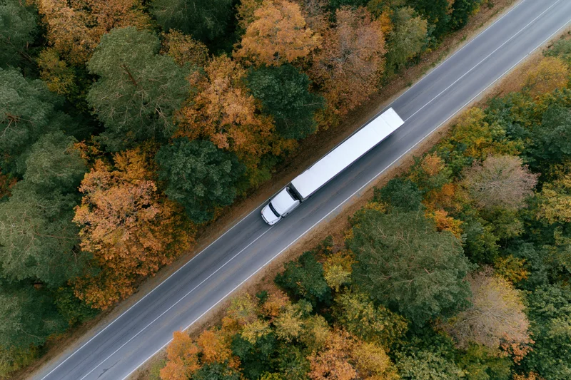 White semi-truck driving on highway through autumn forest from aerial top-down view