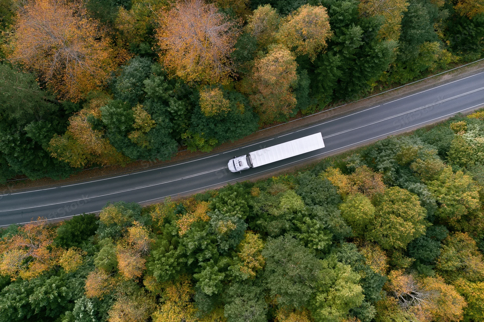 White semi-truck driving on highway through autumn forest from aerial view