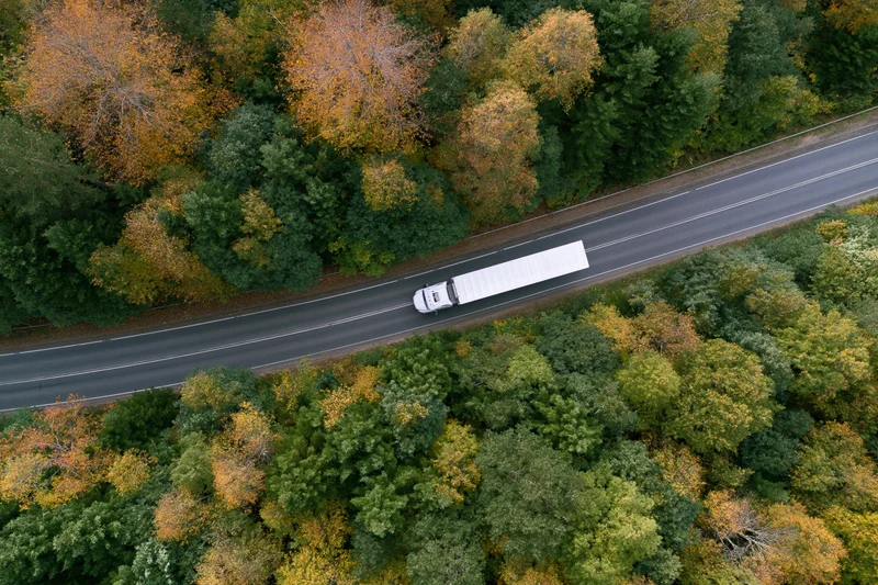 White semi-truck driving on highway through autumn forest from aerial view