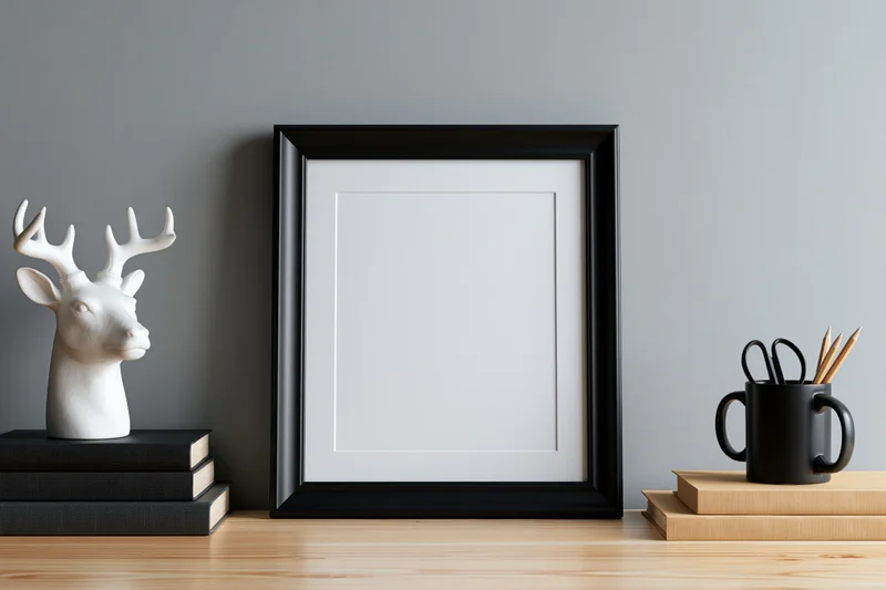 Black picture frame with blank white interior leaning against gray wall on wooden shelf with books, deer statue, and mug holding scissors and pencils