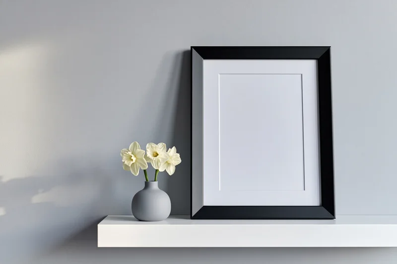 Black picture frame with white mat leaning on white shelf beside vase of daffodils against light gray wall