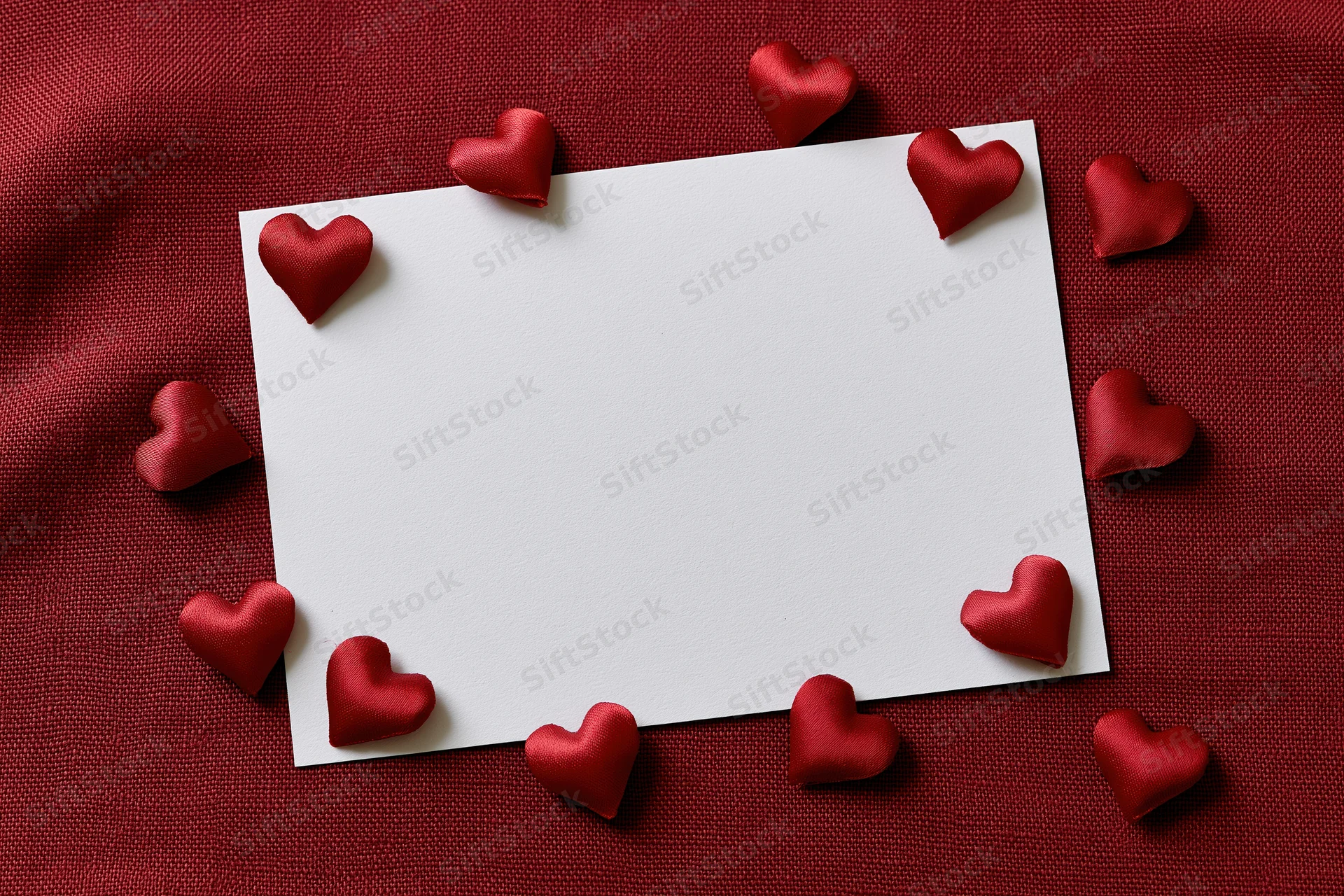 Blank white card surrounded by red satin hearts on red fabric surface